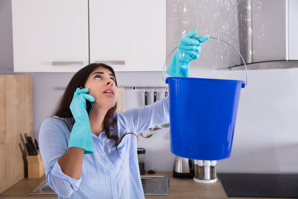 Woman Calling Plumber While Collecting Water Leaking From Ceiling Using Bucket